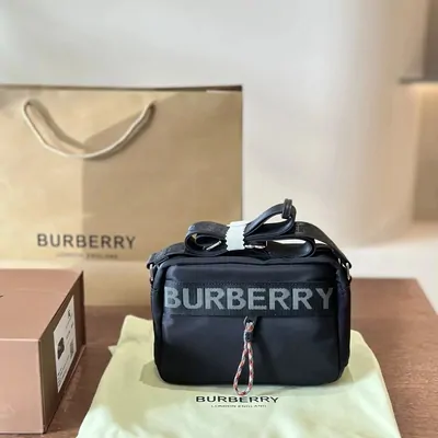 A black nylon crossbody bag with a printed logo, standing on its dust cloth beside a paper carrier bag and a box