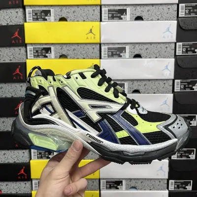 A hand holding up a green, navy and white running shoe in front of a wall of stacked shoe boxes